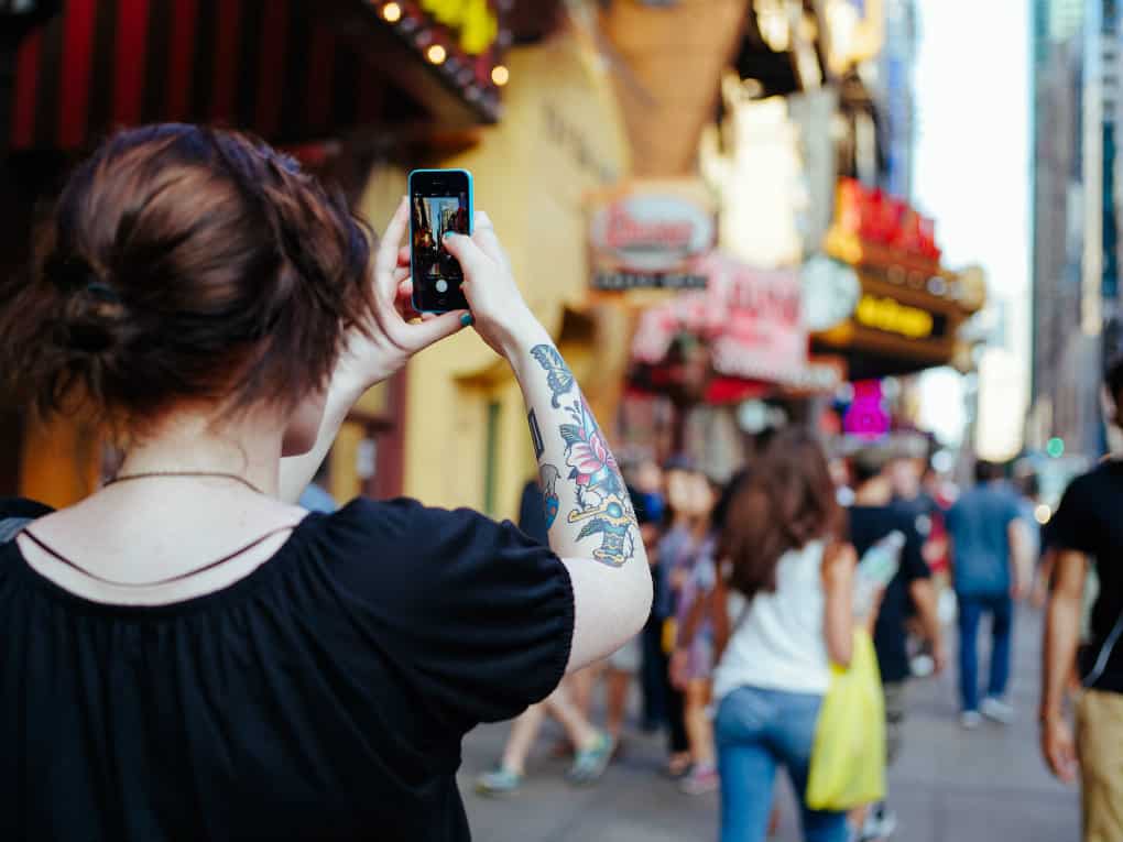 Una ragazza fotografa con lo smartphone una via affollata piena di insegne