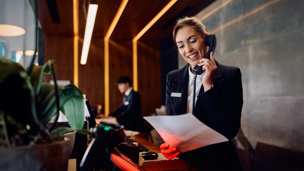 Una receptionist sorridente al telefono legge un foglio al banco di un hotel, con un collega sullo sfondo