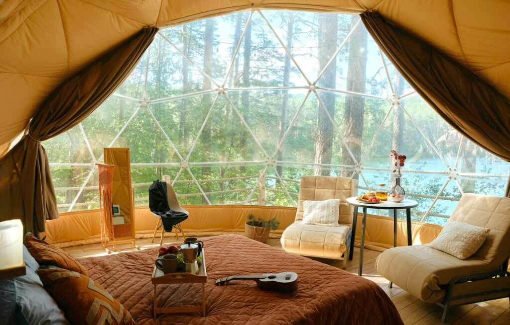 Interno di una tenda a cupola da glamping con letto matrimoniale, poltrone e vetrata sul bosco