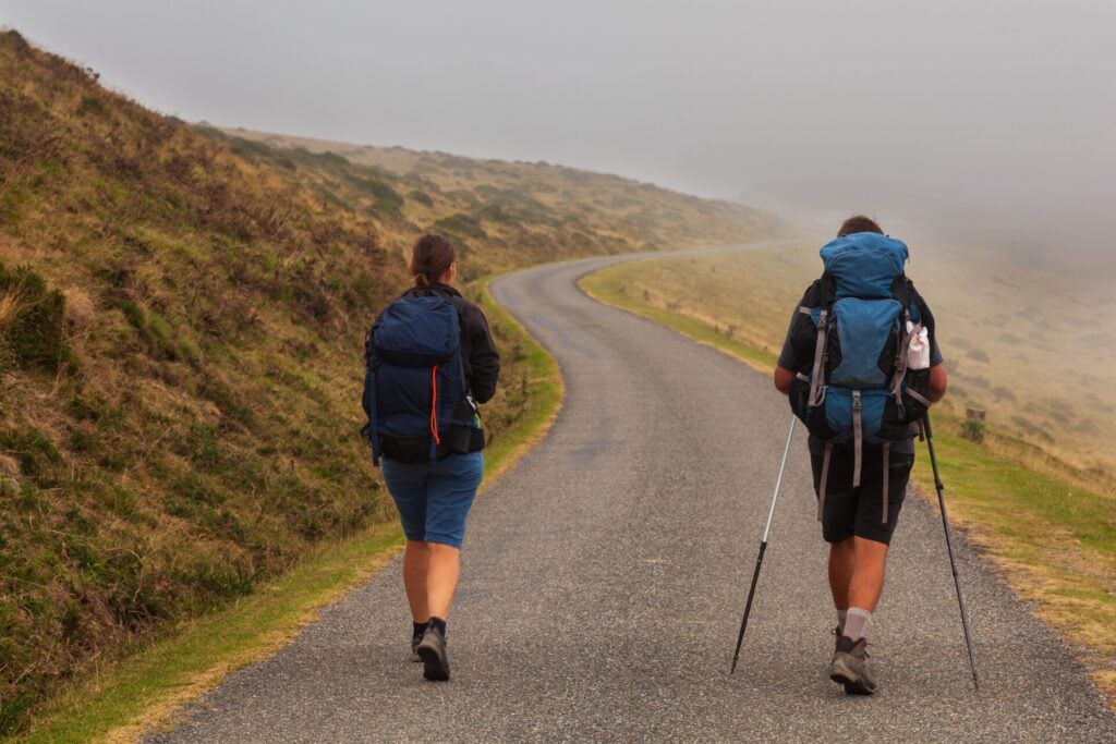 Due escursionisti con zaini e bastoncini camminano su una strada di montagna nella nebbia
