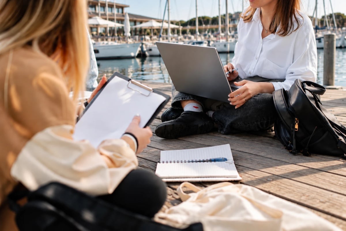 Due studentesse lavorano con portatile, cartellina e quaderno sul pontile di un porto turistico, con le barche a vela alle spalle