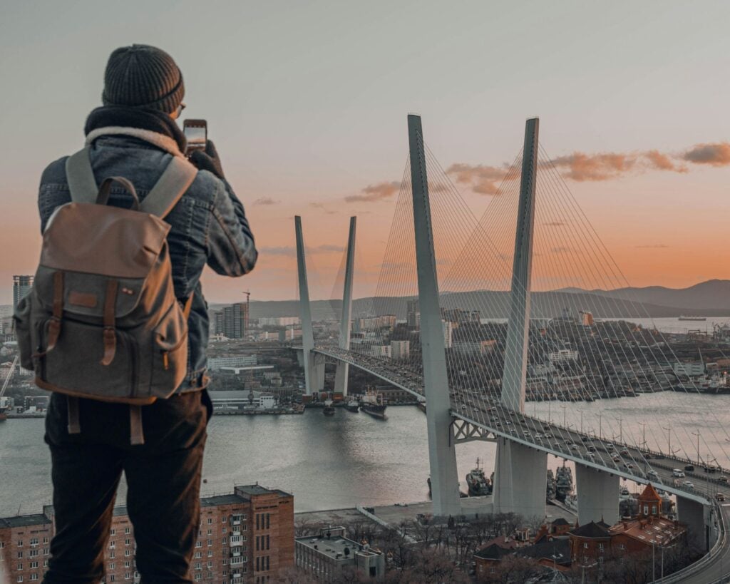 Un ragazzo con lo zaino fotografa con lo smartphone un ponte strallato sulla baia al tramonto